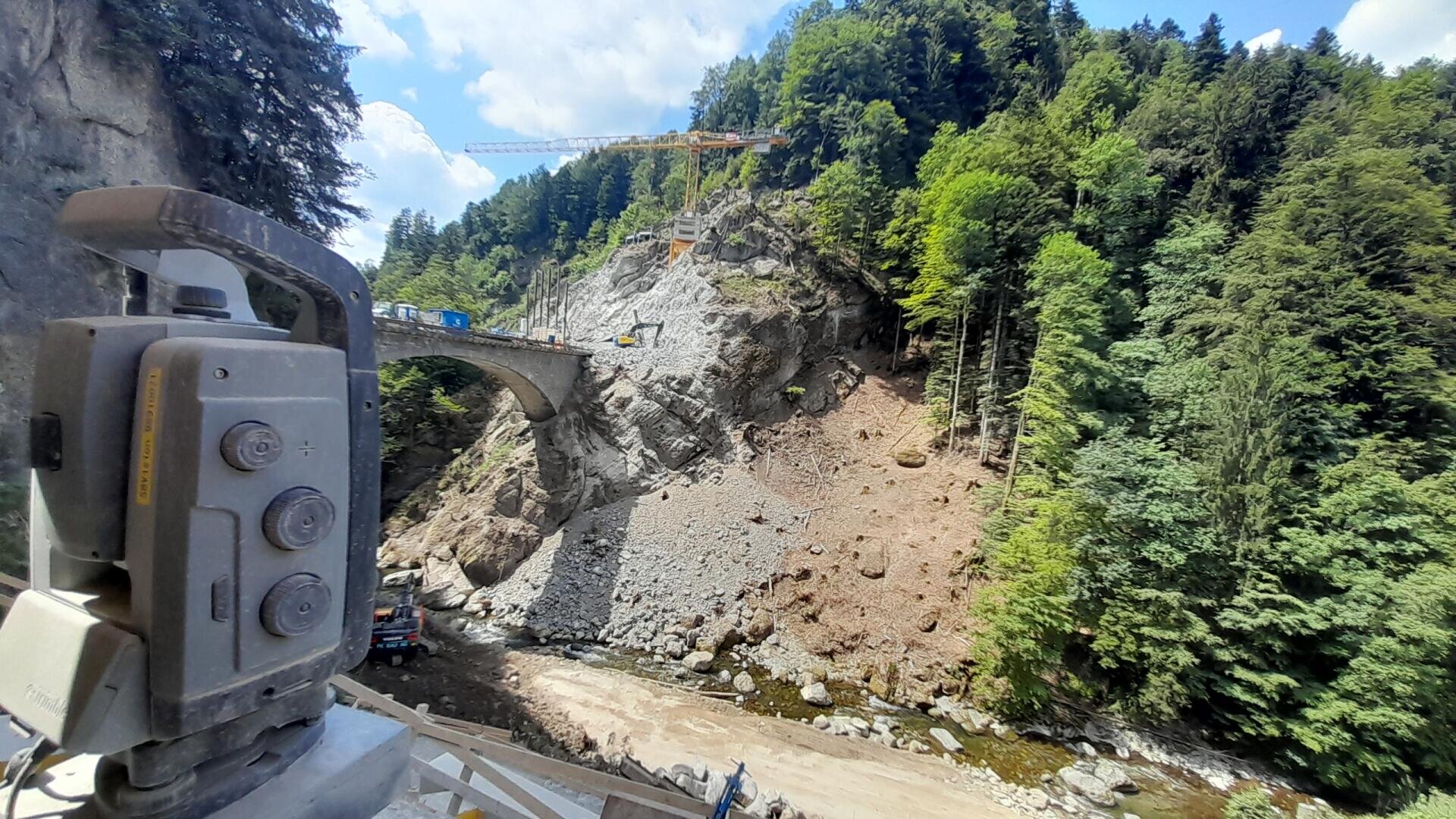 Eine Baustelle in einem bewaldeten Canyon mit einer Brücke, die in einen Felsen gebaut wird, einem Kran darüber und einem Vermessungsinstrument im Vordergrund in der Nähe eines trockenen, felsigen Flussbetts. Helles Tageslicht und blauer Himmel über dem Bild.