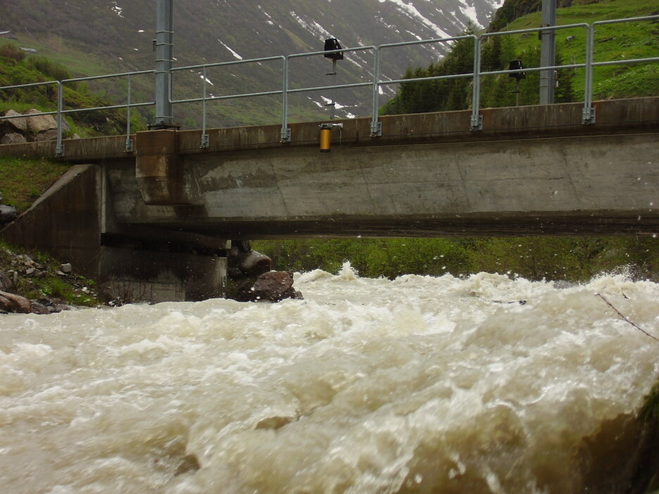 Schnelles, schäumendes Flusswasser fließt unter einer Betonbrücke mit Metallgeländer. Im Hintergrund sind Hänge mit grünem Gras und Schneeflecken zu sehen.