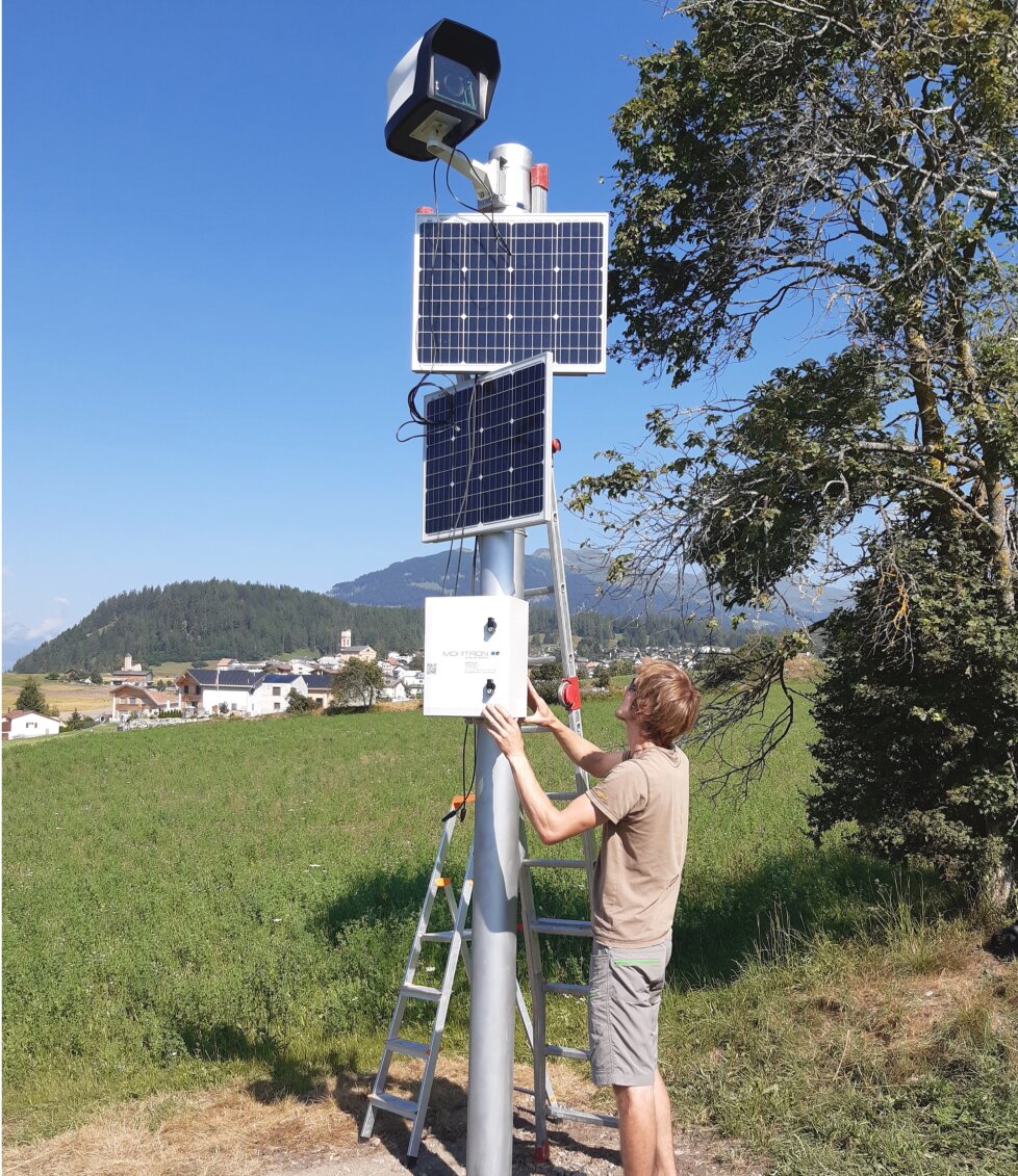 Eine Person steht auf dem Boden neben einer Leiter und arbeitet an einem Metallmast mit Solarmodulen, einer Kamera und Ausrüstungskästen in einem grasbewachsenen Feld mit Hügeln und Gebäuden im Hintergrund.