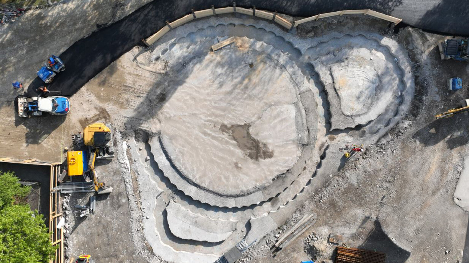 Luftaufnahme eines im Bau befindlichen Skateparks. Zu sehen sind runde und gebogene Betonschalungen, Baufahrzeuge, Arbeiter, Schotter und ein teilweise fertiger Gehweg, der von einem provisorischen Zaun umgeben ist.