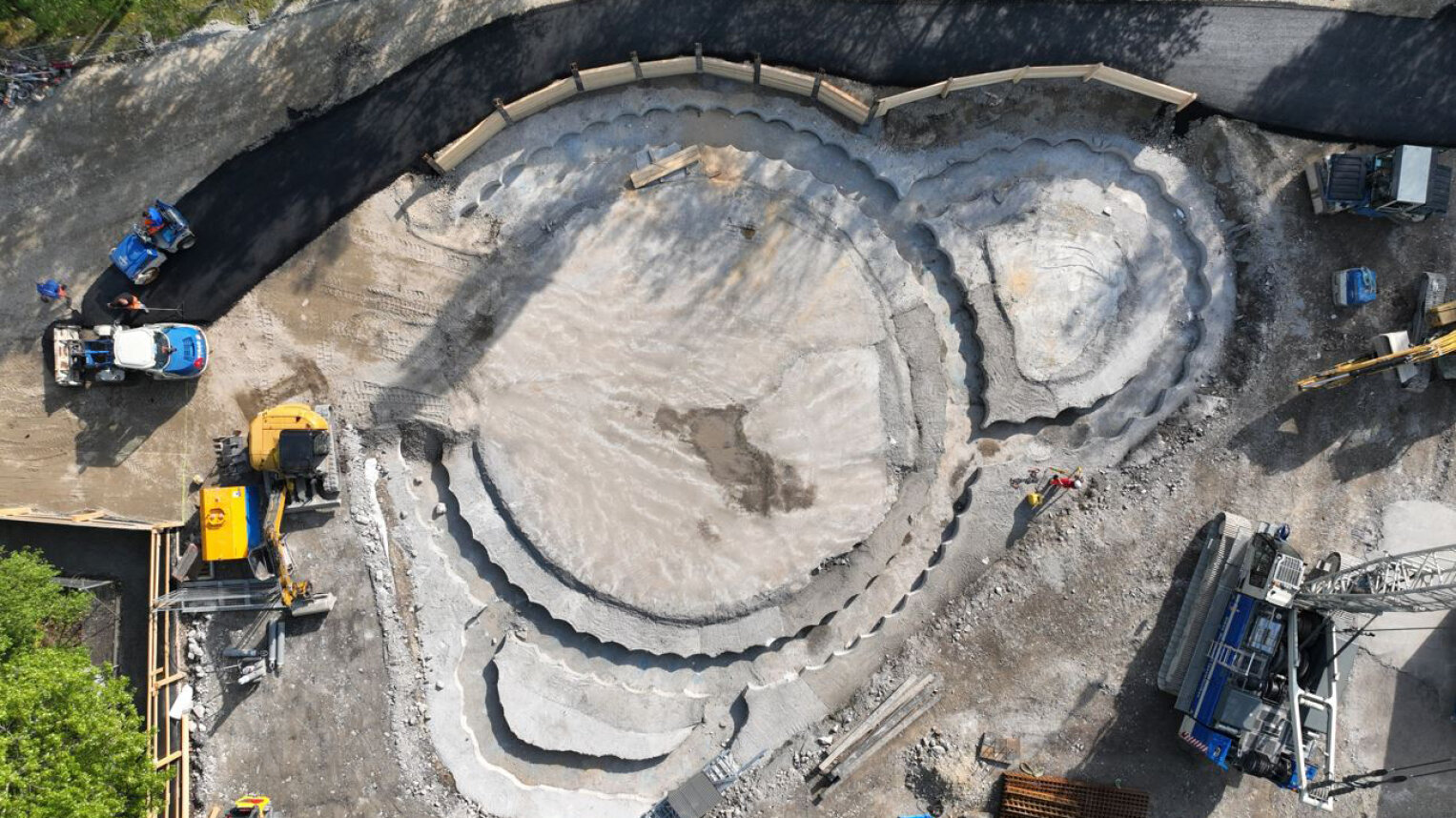 Luftaufnahme einer Baustelle mit einer großen, kreisförmigen Baugrube mit gebogenen, geschichteten Rändern, umgeben von Maschinen, Arbeitern und Baumaterialien. Eine gepflasterte Straße verläuft entlang des oberen Rands.