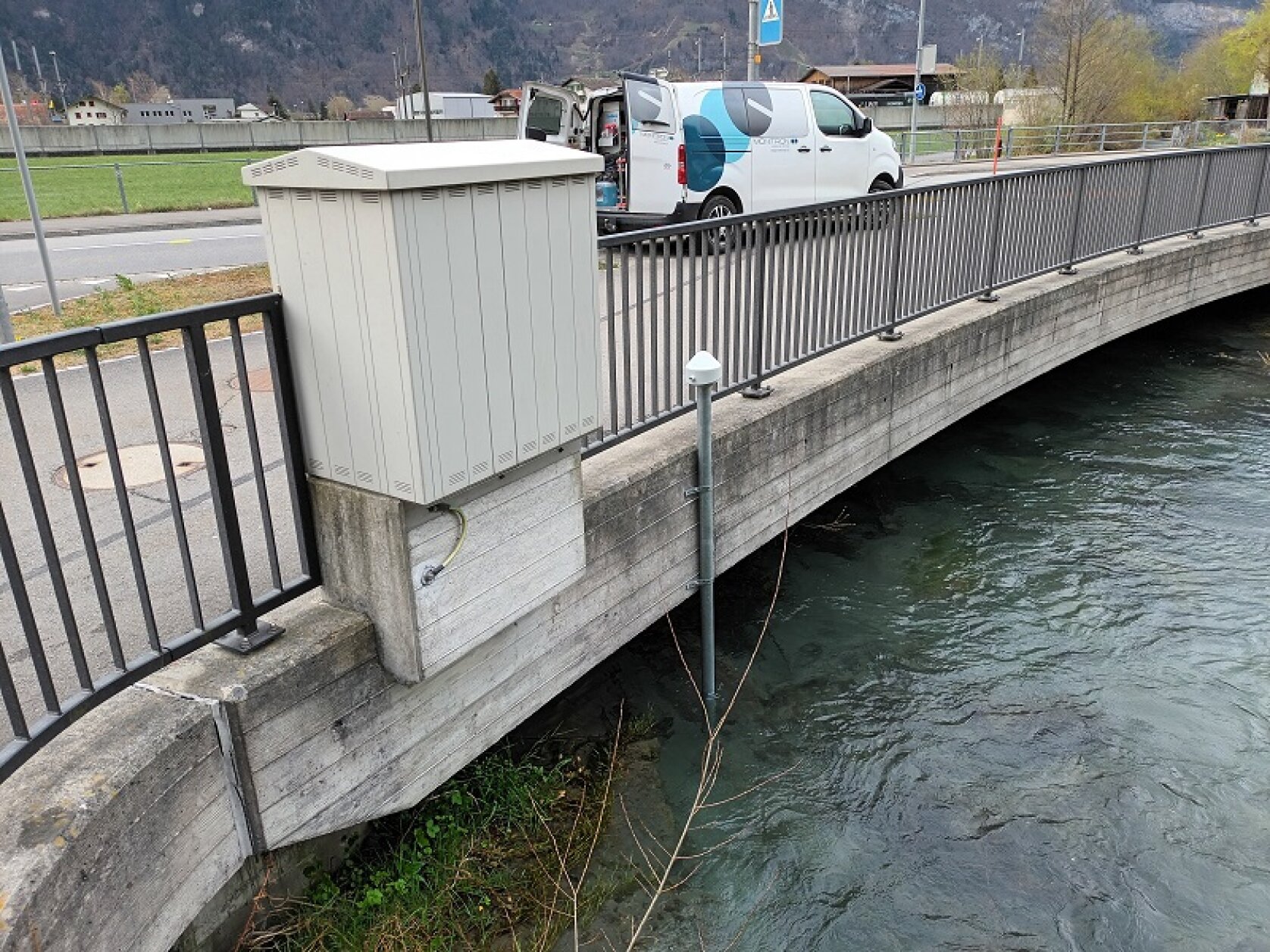 Ein weißer Stromkasten ist an der Seite einer Betonbrücke über einen Fluss angebracht. Unter dem Kasten ist ein Sensor angebracht, der über das Wasser hinausragt. Ein weißer Lieferwagen ist auf der Straße neben der Brücke geparkt.
