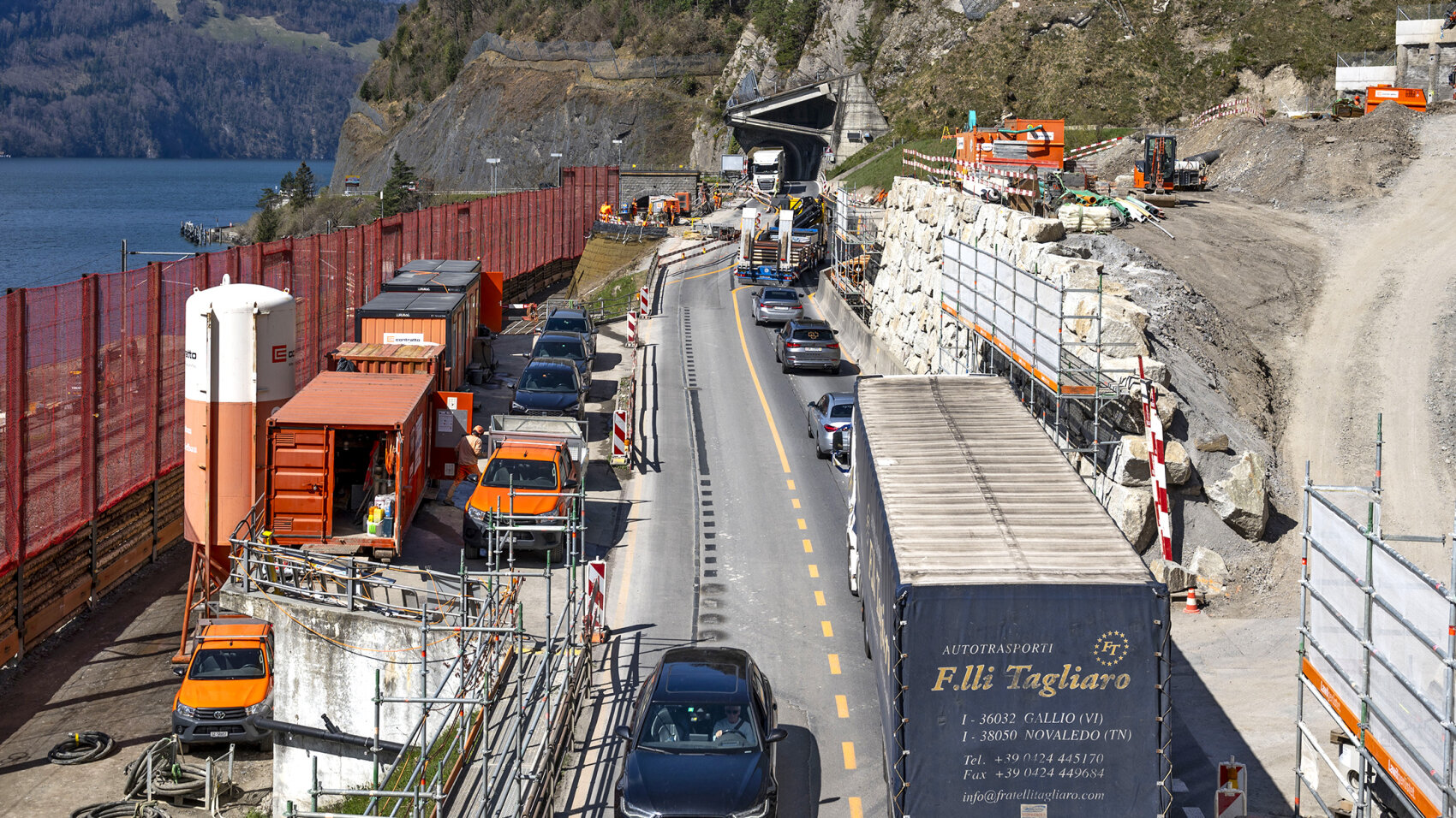 Eine Baustelle auf einer Bergstrasse an einem See, wo sich der Verkehr mit Autos und Lastwagen staut. Orangefarbene Baustellenabsperrungen und -geräte säumen die Strasse, und vor uns ist ein Tunneleingang zu sehen.